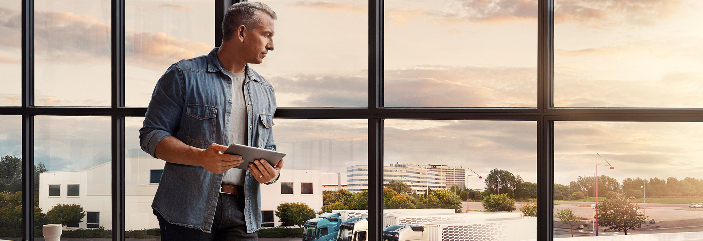 Mężczyzna trzymający tablet stoi przy oknie i spogląda w dół na swoją flotę samochodów ciężarowych